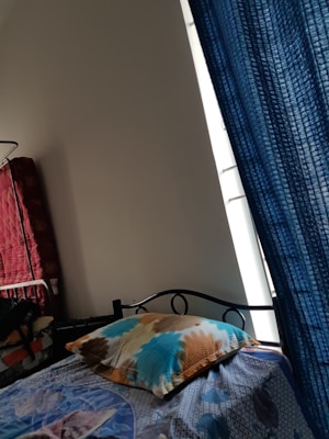 A bed featuring a colorful bedsheet and a pillow is positioned near a window with blue curtains. To the left, there is a red quilted item stacked against the wall, possibly a mattress or blanket. The room has a simple, minimalistic decor with a plain white wall.
