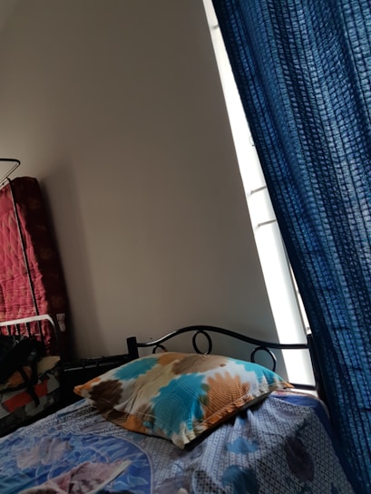 A bed featuring a colorful bedsheet and a pillow is positioned near a window with blue curtains. To the left, there is a red quilted item stacked against the wall, possibly a mattress or blanket. The room has a simple, minimalistic decor with a plain white wall.