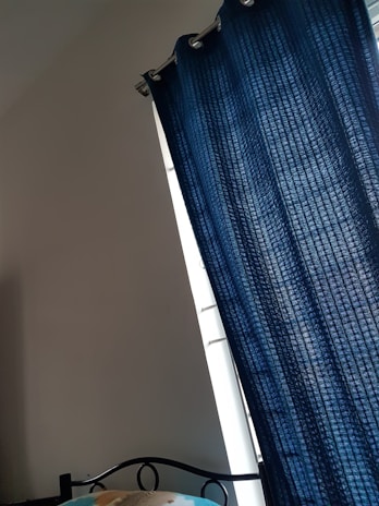 A room corner features a dark blue curtain hanging from a metal rod, partially covering a window. A small portion of a bed with a black metal headboard and a colorful blanket is visible at the bottom.
