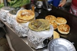 A variety of pancakes or flatbreads sitting on a griddle and wrapped in foil. Some have a golden-brown color while others are darker with different fillings visible. A round metal tray is placed nearby.