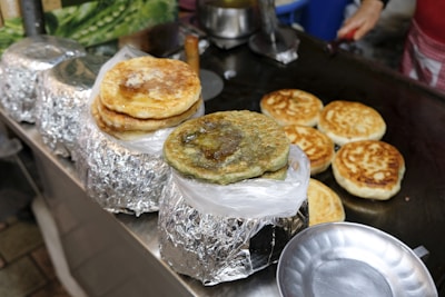 A variety of pancakes or flatbreads sitting on a griddle and wrapped in foil. Some have a golden-brown color while others are darker with different fillings visible. A round metal tray is placed nearby.