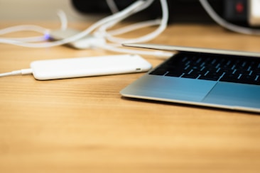 A sleek laptop partially open on a wooden desk, with a portable charger connected via a charging cable next to it.