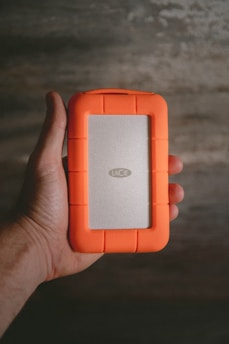 A hand is holding a portable external hard drive with a distinctive bright orange rubber casing and a silver center panel labeled 'LaCie'. The background is a textured surface with a muted color palette, possibly a wooden or concrete surface.