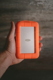A hand is holding a portable external hard drive with a distinctive bright orange rubber casing and a silver center panel labeled 'LaCie'. The background is a textured surface with a muted color palette, possibly a wooden or concrete surface.