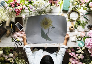 A person is seated at a floral arrangement workspace, holding a sheet with a sunflower drawing. The table is covered with various flowers including tulips, roses, and peonies. Items such as a mobile phone, scissors, ribbons, and twine are scattered around. Green floral foam blocks and tools for crafting are also present.