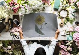 A person is seated at a floral arrangement workspace, holding a sheet with a sunflower drawing. The table is covered with various flowers including tulips, roses, and peonies. Items such as a mobile phone, scissors, ribbons, and twine are scattered around. Green floral foam blocks and tools for crafting are also present.