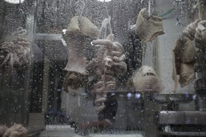 Various cuts of meat, possibly tripe and intestines, are hanging on hooks behind a window covered with water droplets.