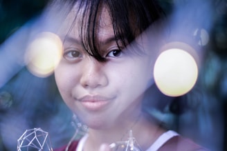 A portrait of a young person with dark hair, slightly smiling, amidst a dreamy, soft-focus background. The image features several out-of-focus lights that create a surreal and artistic atmosphere.