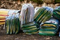 Stacks of colorful magazines are arranged on a surface, with their pages slightly fanned out, creating a layered texture. The setting appears to be outdoors, suggested by the dirt and leaves in the background.