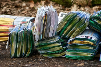 Stacks of colorful magazines are arranged on a surface, with their pages slightly fanned out, creating a layered texture. The setting appears to be outdoors, suggested by the dirt and leaves in the background.