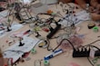 A busy workspace with various electronic components and tools scattered across a light wooden table. Soldering stations, magnifying glasses, wires, pliers, and instruction manuals are visible. Several people are working with the tools, engaging in an electronics project.
