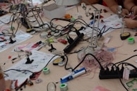 A busy workspace with various electronic components and tools scattered across a light wooden table. Soldering stations, magnifying glasses, wires, pliers, and instruction manuals are visible. Several people are working with the tools, engaging in an electronics project.