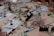 A busy workspace with various electronic components and tools scattered across a light wooden table. Soldering stations, magnifying glasses, wires, pliers, and instruction manuals are visible. Several people are working with the tools, engaging in an electronics project.