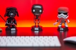 Three Star Wars figurines are lined up on a desk in front of a vibrant red-orange background. The figures include iconic characters from the franchise, each displayed on a black base with the Star Wars logo. A blurred keyboard is visible in the foreground.