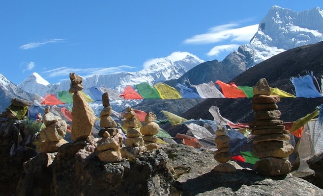 A stunning mountain landscape featuring snow-capped peaks under a clear blue sky. In the foreground, piles of balanced stones are set on a rocky surface. Vibrant, multicolored prayer flags are strung along the scene, waving in the breeze.