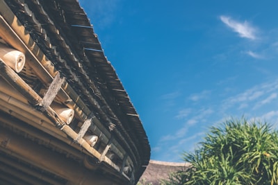 A rustic bamboo roof with wooden supports is prominently featured against a clear blue sky. Lush green foliage is visible in the foreground, adding a touch of nature to the architectural element.