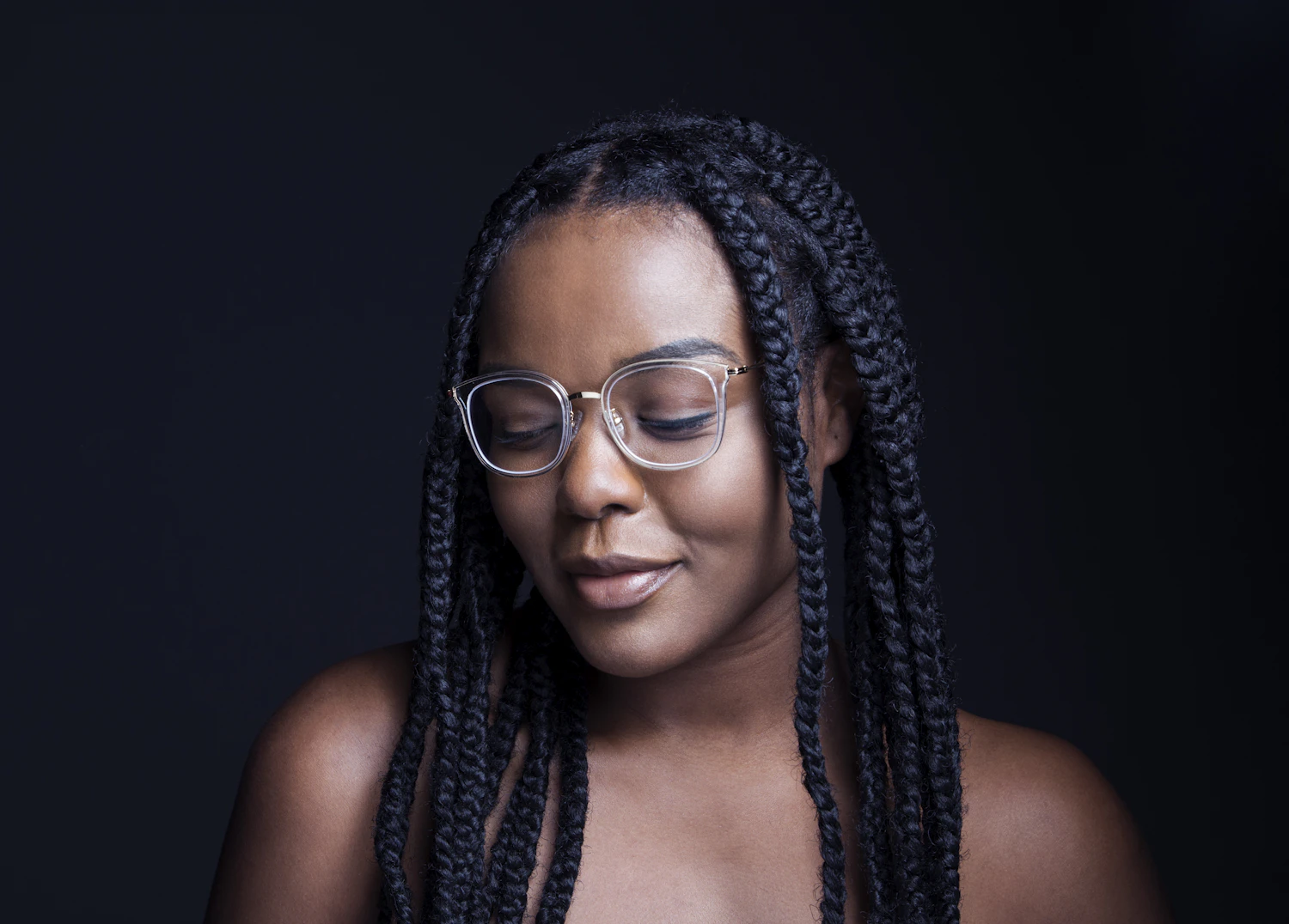 A woman with long braids and pale clear frames photographed against a black background