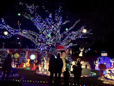 A colorful and festive display of holiday lights adorns a large tree, with vibrant decorations including snowmen, penguins, and various illuminated figures. Silhouettes of people are visible, indicating visitors enjoying the decorations.