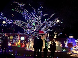 A colorful and festive display of holiday lights adorns a large tree, with vibrant decorations including snowmen, penguins, and various illuminated figures. Silhouettes of people are visible, indicating visitors enjoying the decorations.