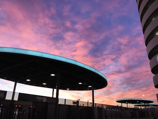 A vibrant sunset with a sky filled with pink and purple hues casts a colorful backdrop behind a modern architectural structure, characterized by circular roofs with lit edges. The silhouette of the building and clear sky gives an urban aesthetic.