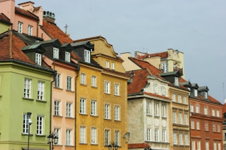 A row of colorful historic buildings with distinct architectural styles, featuring pastel-colored facades and red-tiled roofs. The structures are closely packed and mostly have rectangular windows with white frames. Some buildings have decorative elements such as cornices and ornate window trimmings.