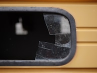 A close-up view of a shattered window with multiple cracks and missing glass pieces. The frame appears to be metallic and is part of a yellow structure, potentially a vehicle or building. The broken glass is dark, with visible fragments and a jagged edge where the glass has been damaged.