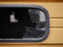 A close-up view of a shattered window with multiple cracks and missing glass pieces. The frame appears to be metallic and is part of a yellow structure, potentially a vehicle or building. The broken glass is dark, with visible fragments and a jagged edge where the glass has been damaged.