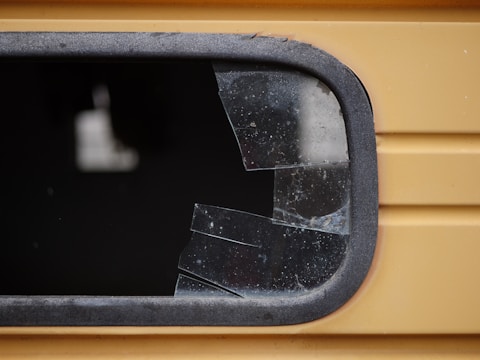 A close-up view of a shattered window with multiple cracks and missing glass pieces. The frame appears to be metallic and is part of a yellow structure, potentially a vehicle or building. The broken glass is dark, with visible fragments and a jagged edge where the glass has been damaged.