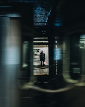A person is standing on a subway platform, visible through the windows of a moving train. The platform is dimly lit, and there is a sign in the background. The scene is framed by the blurred motion of the train, creating a sense of speed and transit.