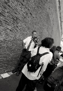 A street musician plays the violin against a textured brick wall. Several people, some with backpacks, stand nearby, possibly listening or passing by. The scene is captured in black and white, adding a classic, timeless feel.