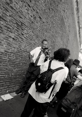 A street musician plays the violin against a textured brick wall. Several people, some with backpacks, stand nearby, possibly listening or passing by. The scene is captured in black and white, adding a classic, timeless feel.