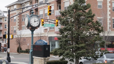 A street scene features a vintage clock on a pole next to a traffic light, with the sign for Palisade Ave visible. In the background, there is a red brick building with white trim and multiple windows, and a visible CVS Pharmacy. A large evergreen tree is in the foreground next to a busy street corner.