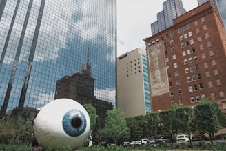 A large, realistic sculpture of a human eye is positioned in a city environment, surrounded by tall buildings with reflective glass windows and brick facades. Lush green trees and shrubs form a backdrop at the bottom of the image, and several vehicles are parked nearby.