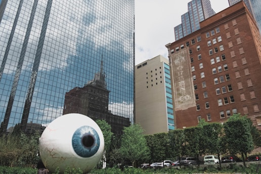 A large, realistic sculpture of a human eye is positioned in a city environment, surrounded by tall buildings with reflective glass windows and brick facades. Lush green trees and shrubs form a backdrop at the bottom of the image, and several vehicles are parked nearby.