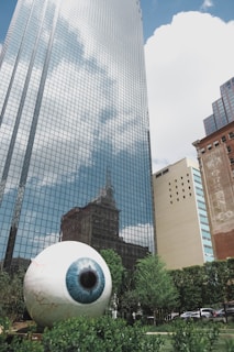 A large, realistic sculpture of an eyeball is situated in a landscaped area with greenery in the foreground. Behind the sculpture, there's a towering modern glass building reflecting the sky and surrounding architecture. Adjacent to it are additional buildings of various architectural styles.