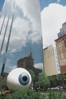 A large, realistic sculpture of an eyeball is situated in a landscaped area with greenery in the foreground. Behind the sculpture, there's a towering modern glass building reflecting the sky and surrounding architecture. Adjacent to it are additional buildings of various architectural styles.
