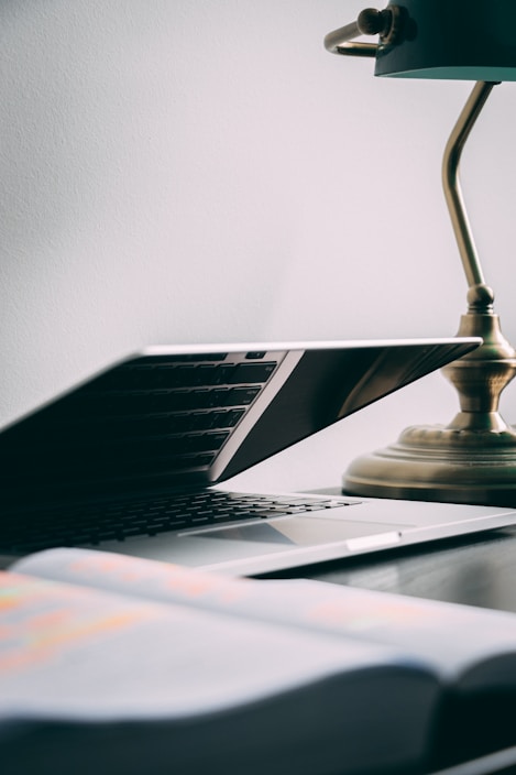 A partially opened laptop is placed on a dark wooden desk alongside a brass desk lamp. In the foreground, there is a blurred book or notebook. The scene is minimalist and well-lit by natural light, casting shadows and reflections on the laptop.