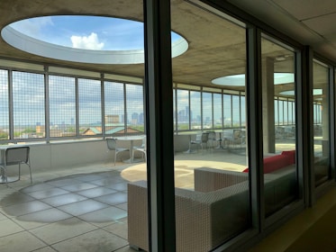 A modern rooftop terrace with large circular openings in the ceiling and metal grid walls, providing a panoramic city view. The area is furnished with metallic chairs and tables, and features a red cushioned sofa. The terrace is partially indoors, seen through glass windows, with sunlight casting geometric shadows onto the tiled floor.