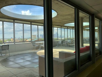 A modern rooftop terrace with large circular openings in the ceiling and metal grid walls, providing a panoramic city view. The area is furnished with metallic chairs and tables, and features a red cushioned sofa. The terrace is partially indoors, seen through glass windows, with sunlight casting geometric shadows onto the tiled floor.