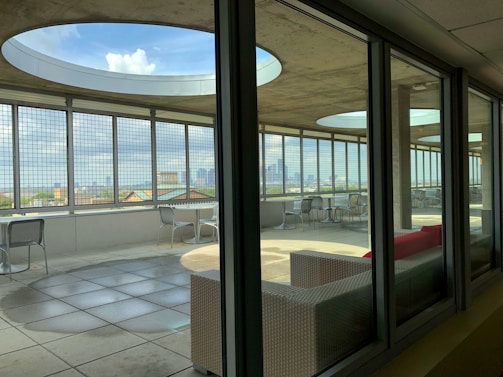 A modern rooftop terrace with large circular openings in the ceiling and metal grid walls, providing a panoramic city view. The area is furnished with metallic chairs and tables, and features a red cushioned sofa. The terrace is partially indoors, seen through glass windows, with sunlight casting geometric shadows onto the tiled floor.