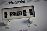 A close-up view of a white Hotpoint appliance control panel, featuring a mechanical timer display indicating 2.20. Two white knobs labeled 'Minute Minder' and 'Cook Time/Start Time' are present below the display.