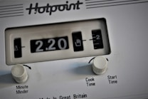 A close-up view of a white Hotpoint appliance control panel, featuring a mechanical timer display indicating 2.20. Two white knobs labeled 'Minute Minder' and 'Cook Time/Start Time' are present below the display.
