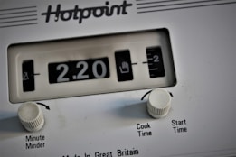 A close-up view of a white Hotpoint appliance control panel, featuring a mechanical timer display indicating 2.20. Two white knobs labeled 'Minute Minder' and 'Cook Time/Start Time' are present below the display.