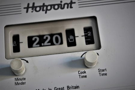 A close-up view of a white Hotpoint appliance control panel, featuring a mechanical timer display indicating 2.20. Two white knobs labeled 'Minute Minder' and 'Cook Time/Start Time' are present below the display.