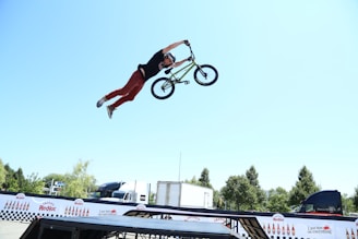 A person performs an aerial stunt on a BMX bike, extending their body horizontally with one hand gripping the handlebars. The scene is set outdoors with a bright blue sky and trees in the background. Various vehicles and promotional banners are visible, adding an event-like atmosphere.