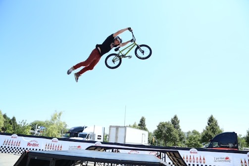 A person performs an aerial stunt on a BMX bike, extending their body horizontally with one hand gripping the handlebars. The scene is set outdoors with a bright blue sky and trees in the background. Various vehicles and promotional banners are visible, adding an event-like atmosphere.