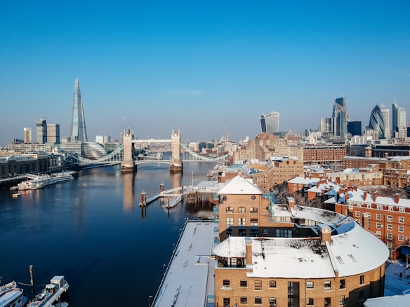 A wintery cityscape of London featuring the iconic Tower Bridge and the Shard. The river Thames runs through the scene, reflecting the clear blue sky. Snow lightly covers the rooftops of the surrounding buildings, creating a picturesque contrast against the brick structures. Modern skyscrapers are visible in the background.