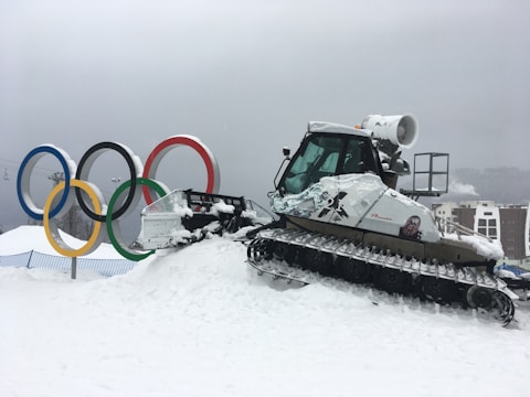 A snow-covered plowing vehicle is parked on a slope in a winter setting. In the background, there are large, colorful Olympic rings, and the scene is overcast, suggesting cloudy weather. Buildings are partially visible, indicating a developed area nearby.
