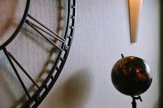 A large, ornate metal clock with Roman numerals is partially visible on the left side. To the right of the clock, there is a dark-colored globe with continents in shades of brown and gold. The globe is mounted on a stand. Above the globe, there is a modern, vertical wall sconce emitting a soft light. The background is a textured, light-colored wall.