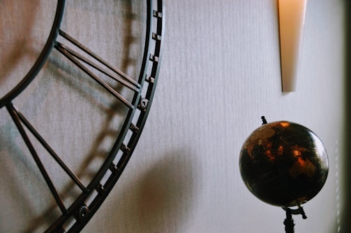 A large, ornate metal clock with Roman numerals is partially visible on the left side. To the right of the clock, there is a dark-colored globe with continents in shades of brown and gold. The globe is mounted on a stand. Above the globe, there is a modern, vertical wall sconce emitting a soft light. The background is a textured, light-colored wall.